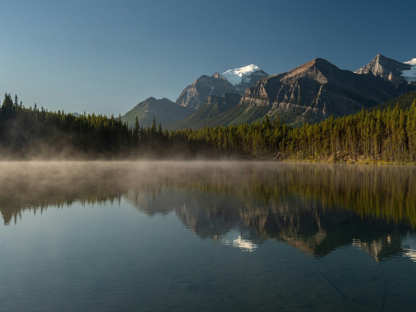 Mist rises from a lake nestled among snow capped mountains.