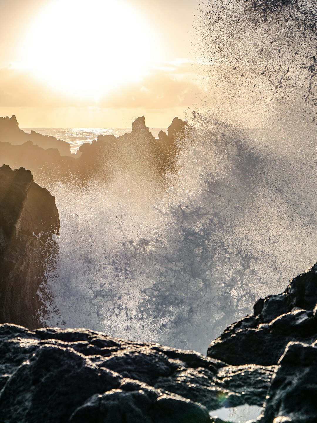 Ocean waves crash against dark jagged rocks at sunset.